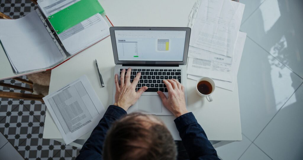 Top-down wide shot of man’s hands on laptop at home, calculating and summarizing his taxes in excel with cup of coffee next to the laptop and large number of documents scattered around the table