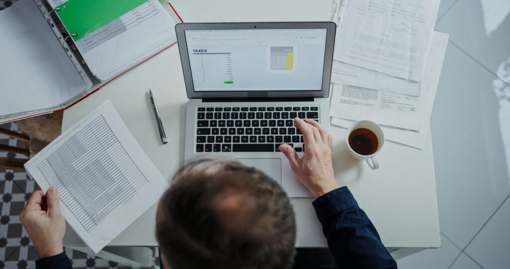 Top-down wide shot of a man’s hands on a laptop at home, working on his taxes in Excel and sipping coffee, with many documents spread across the table